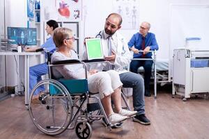 Doctor with green screen tablet in rehabilitation center for disabled patients. Man with disabilities ,walking frame sitting in hospital bed. Health care system, clinic patients. photo