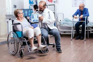 Doctor showing brain X ray image of disabled elderly senior woman in wheelchair. Man with disabilities ,walking frame sitting in hospital bed. Health care system, clinic patients. photo