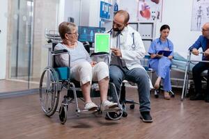 Doctor pointing at green mockup on tablet pc while telling senior woman in wheelchair about her rehabilitation. Man with disabilities ,walking frame sitting in hospital bed. Health care system, photo