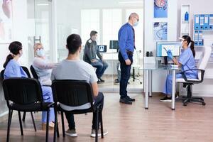 Hospital waiting room with patients sitting on chairs and nurse explaining treatment to elderly man wearing face mask against covid-19. Social distancing, medic, covid-19, man, worried, protection photo