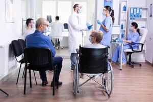 Disabled senior woman wearing face mask against coronavirus in hospital waiting area and having a conversation with old man. Assistant working on reception computer. photo