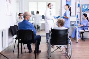 Doctor discussing with nurse in hospital waiting area and disabled senior woman in wheelchair having a conversation with old man. Assistant working on reception computer. photo