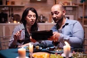 alegre joven Pareja comida juntos utilizando tableta durante romántico cena. adultos sentado a el mesa en el cocina hojeada, buscando, , Internet, celebrando, aniversario, feliz., festivo, disfrutar. foto