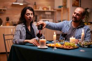 novio celebrando relación con esposa en comida habitación. joven hombre torrencial rojo vino en esposa vaso. romántico caucásico contento Pareja sentado a el mesa celebrando . foto