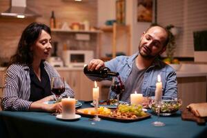 marido torrencial esposa a esposa durante romántico cena en comida habitación. joven hombre torrencial rojo vino en esposa vaso. romántico caucásico contento Pareja sentado a el mesa celebrando . foto