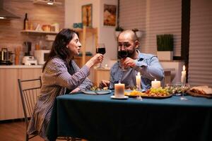 Pareja tostado con vino lentes durante romántico cena. relajarse contento personas cantarín, sentado a mesa en cocina, disfrutando el comida, celebrando aniversario en el comida habitación. foto