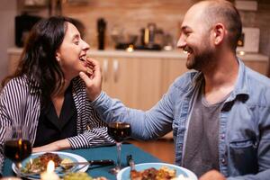 Husband feeding wife with grapes during romantic dinner celebrating relationship. Wife and husband celebrating anniversary with red wine, tender moments at candle lights in kitchen. photo