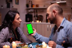 Guy holding phone with green screen while having romantic dinner with wife. Happy looking at green screen template chroma key isolated smartphone display using technology internet. photo