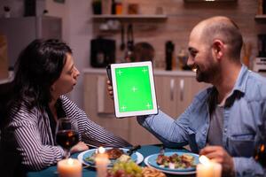 Couple using tablet pc with chroma key and enjoying their time together during romantic dinner. Husband and wife looking at green screen template chroma key display sitting at the table in kitchen during dinner. photo