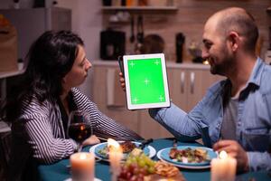 Husband holding tablet pc with green mock up while enjoying romantic dinner with wife. Man and woman looking at green screen template chroma key display sitting at the table in kitchen during dinner. photo