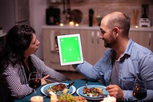 Pareja utilizando tableta ordenador personal con verde modelo durante festivo cena. marido y esposa mirando a verde pantalla modelo croma llave monitor sentado a el mesa en cocina. foto