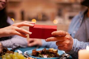 Close up of gift box holded by husband and wife during romantic dinner. Cheerful man dining with woman at home, enjoying the meal, celebrating their anniversary at candle lights photo