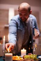 Husband lighting candle on kitchen table a for festive dinner with wife. Man preparing festive meal with healthy food for anniversary celebration, romantic date. photo