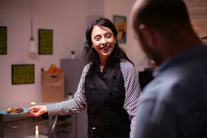 Wife smiling at husband wearing apron while looking at husband after cooking tasty dinner. Woman cooking festive food for her and husband to celebrate relationship and romance, happiness, positive, love, emotion, marriage. photo