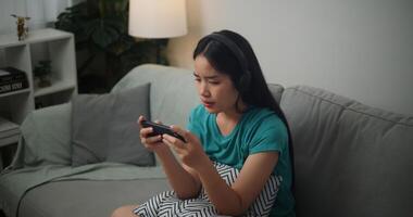 retrato de joven asiático mujer vistiendo lentes y auriculares disfruta jugando en línea deporte juegos en teléfono inteligente sentado en sofá en el vivo habitación a hogar, jugador estilo de vida concepto. foto