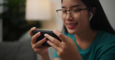 retrato cerca arriba de joven asiático mujer vistiendo lentes y auriculares disfruta jugando en línea deporte juegos en teléfono inteligente sentado en sofá en el vivo habitación a hogar, jugador estilo de vida concepto. foto