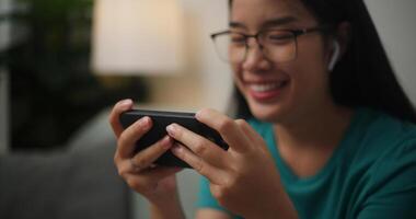 retrato cerca arriba de joven asiático mujer vistiendo lentes y auriculares disfruta jugando en línea deporte juegos en teléfono inteligente sentado en sofá en el vivo habitación a hogar, jugador estilo de vida concepto. foto