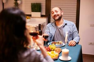 marido sensación contento sonriente a esposa durante romántico cena. hablando contento sentado a mesa comida habitación, disfrutando el comida a hogar teniendo romántico hora a vela luces. foto