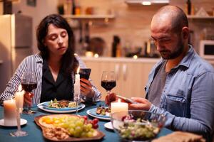 Couple holding glasses of wine and using smartphone in kitchen during romantic dinner with candles and food in the table. Adults sitting at the table browsing, searching, using smartphones, internet, photo