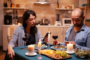 Happy couple watching video on smartphone in kitchen during anniversary. Adults sitting at the table in the kitchen browsing, searching, using smartphones, internet, celebrating anniversary. photo