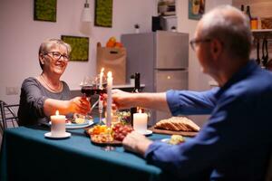 Happy pensioner couple toasting with red wine glasses in kitchen. Senior couple sitting at the table in kitchen, talking, enjoying the meal, celebrating their anniversary in the dining room. photo