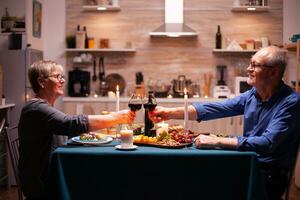 mayor Pareja tostado lentes con rojo vino durante cena. mayor hombre y mujer sentado a el mesa en cocina, hablando, disfrutando el comida, celebrando su aniversario en el comida habitación. foto