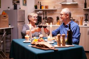 Elderly couple toasting wine glasses for festive dinner. Happy cheerful senior elderly couple dining together in the cozy kitchen, enjoying the meal, celebrating their anniversary. photo