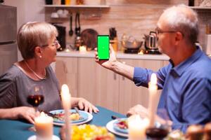 Mature man using phone with green screen in kitchen during romantic dinner with wife. Aged people looking at mockup template chroma key isolated smart phone display using techology internet. photo