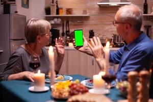 mayor hombre y mujer ondulación a teléfono con verde pantalla. Envejecido personas mirando a Bosquejo modelo croma llave aislado inteligente teléfono monitor utilizando tecnología Internet sentado a el mesa en cocina. foto