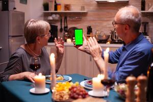 Aged man holding phone with green screen and wife is waving at it. Aged people looking at mockup template chroma key isolated smart phone display using techology internet sitting at the table photo