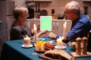 verde pantalla tableta a cena. Envejecido personas mirando a verde pantalla modelo croma llave monitor sentado a el mesa en cocina durante cena. foto