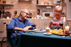 Grandmother looking at gift from disabled husband in wheelchair. Happy cheerful elderly couple dining together at home, enjoying the meal, celebrating their marriage. photo