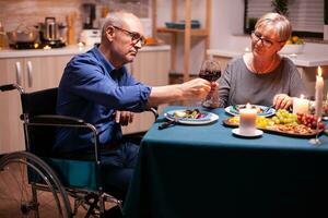 Man with disabilities having dinner with wife and clinking wine glass. Wheelchair immobilized paralyzed handicapped man dining with wife at home, enjoying the meal photo