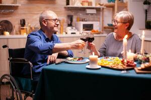 Disabled man smiling at wife while having dinner in kitchen. Wheelchair immobilized paralyzed handicapped man dining with wife at home, enjoying the meal. photo