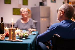 Handicapped man holding whine glass sitting in wheelchair during festive dinner. Happy cheerful senior elderly couple dining together in the cozy kitchen, enjoying the meal, celebrating their anniversary. photo