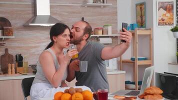 contento joven Pareja tomando selfies durante desayuno. alegre casado marido y esposa haciendo gracioso caras mientras tomando un foto durante desayuno en cocina.