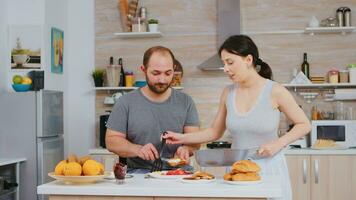 esposa Cocinando huevos para marido durante desayuno mientras él es manchando mantequilla en asado un pan. vistiendo pijama en el mañana, preparando comida juntos, joven contento Pareja amor y matrimonio foto