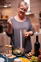 Wife looking at burning candle while preparing table for romantic dinner with husband. Elderly woman waiting her husband for a romantic dinner. Mature wife preparing festive meal for anniversary celebration. photo