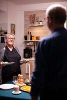 Romantic senior wife in kitchen talking with husband while serving romantic dinner. Elderly old couple talking, sitting at the table in kitchen, enjoying the meal, celebrating their anniversary . photo