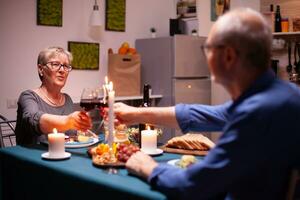 Senior couple clinking glasses with red wine during evening in kitchen. Elderly couple sitting at the table in kitchen, talking, enjoying the meal, celebrating their anniversary in the dining room. photo