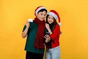 alegre joven asiático Pareja vistiendo Papa Noel sombrero suéteres utilizando crédito tarjeta y teléfono inteligente aislado en amarillo antecedentes. foto