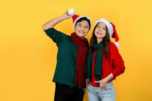 Happy Asian couple in bright green and red outfits and Christmas hats smiling at the camera on a yellow background. photo