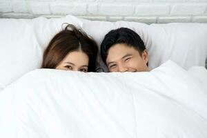 Asian couple under duvet looking at camera on white bed photo