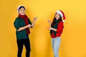 Attractive young Asian man and woman wearing Santa hats pointing at copy space on yellow background. photo