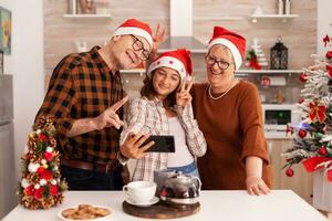 Happy family taking selfie using smartphone making funny expressions during photo time enjoying christmas holiday together in xmas decorated culinary kitchen. Grandchild celebrating winter season