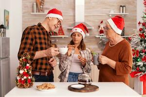 Smiling family celebrating christmas season together in xmas decorated culinary kitchen eating delicious cookies, drinking coffee enjoying winter holiday. Grandchild having fun during christmastime photo