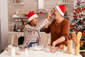 Happy family enjoying winter season putting flour on grandma nose while preparing homemade gingerbread dough in xmas decorated kitchen. Child cooking traditional cookies celebrating christmas holiday photo