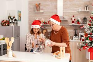 familia celebrando Navidad fiesta preparando hecho en casa masa postre en Navidad decorado culinario cocina a hogar. abuela poniendo harina en colador mientras niño sacudir disfrutando invierno temporada foto