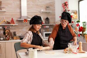 Happy family with apron preparing homemade dough cooking xmas gingerbread dessert in x-mas decorated kitchen. Granddaughter using baking rolling pin making delicious cookies enjoying christmas season photo