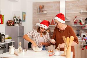 Grandchild mixing ingredients in bowl preparing traditional homemade dough with grandmother celebrating christmas season together in xmas decorated kitchen. Kid enjoying winter holiday baking cookies photo